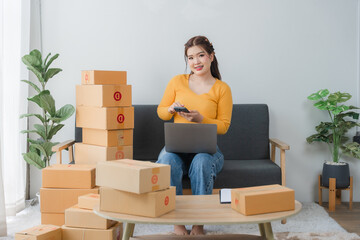 E-commerce Hustle: Young Asian entrepreneur managing orders and logistics on phone and tablet, surrounded by shipping boxes in her home office.	