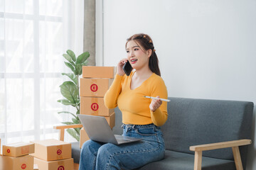 E-commerce Hustle: Young Asian entrepreneur managing orders and logistics on phone and tablet, surrounded by shipping boxes in her home office.	