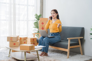 E-commerce Hustle: Young Asian entrepreneur managing orders and logistics on phone and tablet, surrounded by shipping boxes in her home office.	