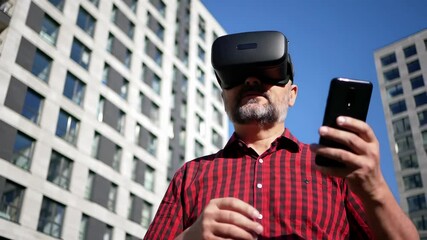 Man using virtual reality headset with smartphone outdoors in urban environment. Modern technology experience with vr device and mobile phone in contemporary city setting with office buildings. - Powered by Adobe
