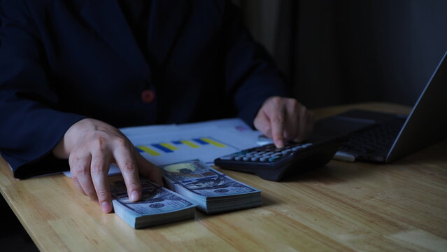 Ideal for content related to economics, money management, profit calculation, business development. holds U.S. dollar bills while working with a calculator and financial graph papers on a table.