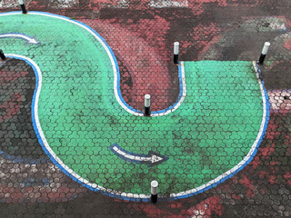 A painted, winding green path on a textured, multi-colored pavement, marked with white arrows.