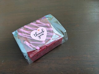 A single red velvet brownie, individually wrapped in cellophane, adorned with pink icing and a...
