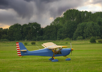 Blue and yellow sport airplane on a grassy airfield beneath dramatic storm clouds. Ideal for...