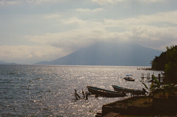 Scenic view of Lake Atitlán with still waters and a distant volcano. Boats are docked along the shoreline, highlighting a serene and peaceful natural landscape during a sunny day. Taken on 35mm Film.