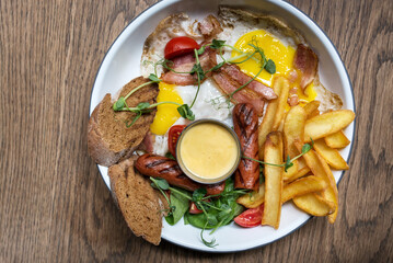 Traditional full English breakfast with fried eggs, sausages, bacon, toast, golden fries, sauce, and fresh vegetables. Served on a white plate on a rustic wooden table
