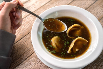 Hot clear dark broth with dumplings, sprinkled with fresh dill, served in a white bowl on rustic wooden table. A hand holding a spoon with soup above the bowl