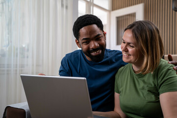 Happy diverse couple smiling and looking at laptop in bright living room during daytime, enjoying love and positive mood.