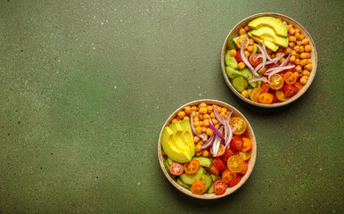 Two bowls of healthy chickpea salad with ripe avocado, juicy cherry tomatoes, crisp cucumber, and red onion slices. Perfect for a nutritious and healthy meal on green background, copy space