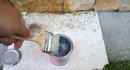 A hand dips a brush into a metal can of solution, preparing for work near a stone wall. The...