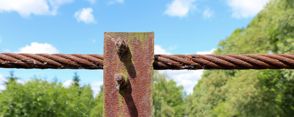 A gloomy day emphasizes the texture of rust on a thick metal cable, secured to a post. This creates...