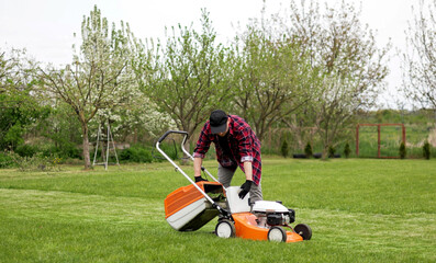 Man in casual clothes takes out bag with cut grass of lawn mower after mowing lawn with at own...