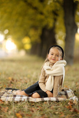 Adorable little child girl 6-7 years old smiling looks at camera, sits on plaid at outdoors at autumn park. Children's day. Stylish kid wears beige sweater and black headband. Happy childhood.