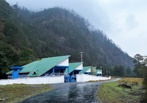 A beautiful country road passing by a house with inclined roof in green color with a view of green hills in the backdrop, image of dong resort in arunachal pradesh