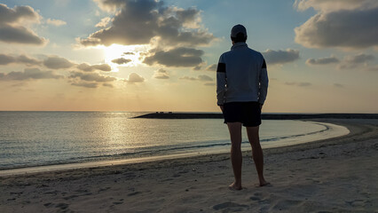 A lone man in a cap stands on a sandy beach in Okinawa, watching the beautiful golden sunset over the tranquil ocean, creating a scene of peace, solitude, and contemplation during a coastal vacation.