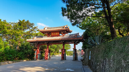 Iconic red Shureimon Gate of Shuri Castle stands majestically under clear blue sky in Naha, Okinawa, its traditional Ryukyuan architecture welcoming visitors to historic UNESCO World Heritage site.