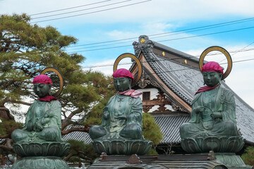 Three serene bronze statues of Jizo Bodhisattvas, adorned with traditional red bibs and caps, sit...
