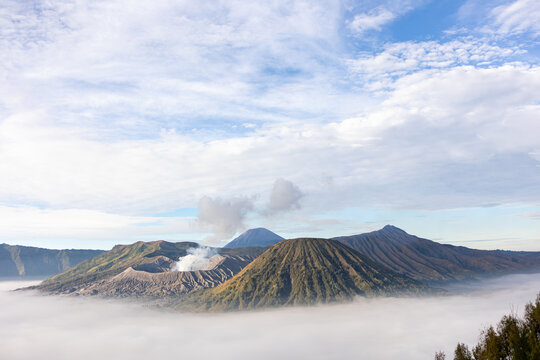 View of majestic volcanoes rising above a sea of ethereal mist under a vast sky with scattered clouds, Mount Bromo Volcano, East Java, Indonesia.