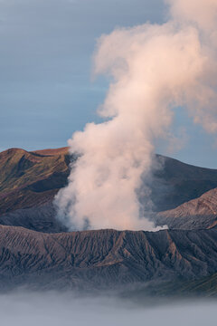 View of a volcanic landscape with wisps of white smoke rising into the blue sky, contrasting against the dark, rugged terrain, Mount Bromo, East Java, Indonesia.