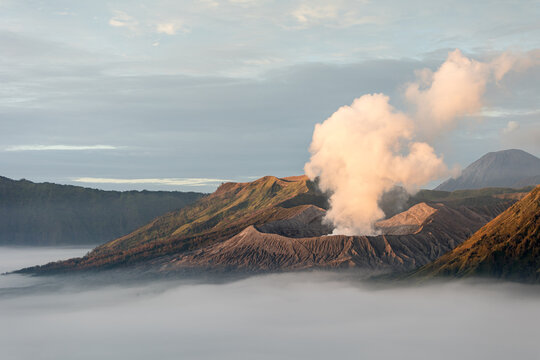 View of the Mount Bromo Volcano erupting with smoke, surrounded by a sea of clouds, creating a dramatic and ethereal landscape, Mount Bromo, East Java, Indonesia.