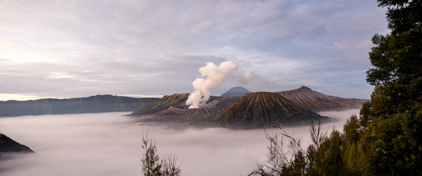 View of the majestic Mount Bromo volcano piercing through a sea of white clouds, with plumes of smoke rising into the sky, Mount Bromo Volcano, East Java, Indonesia.