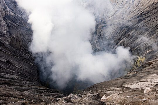 View of plumes of white smoke billowing from the depths of a volcanic crater, a stark contrast against the dark, rugged terrain, Mount Bromo, East Java, Indonesia.