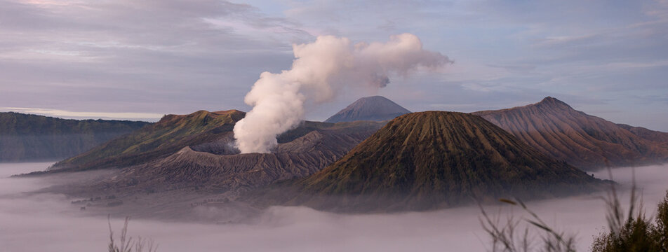 View of majestic volcanoes piercing through a sea of clouds, with white smoke billowing from the crater, Mount Bromo Volcano, East Java, Indonesia.