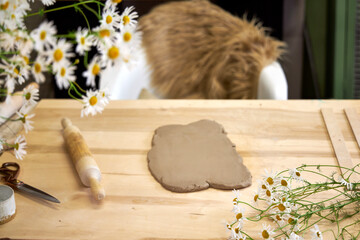 Clay workshop setup with rolled-out clay, rolling pin, tools, and fresh chamomile flowers on a...