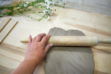 Close-up of a hand rolling out clay on a wooden surface with a rolling pin. Chamomile flowers in...
