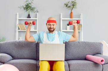 Happy young man in retro sportswear with dumbbells looking at laptop. Bearded funny guy sitting on...