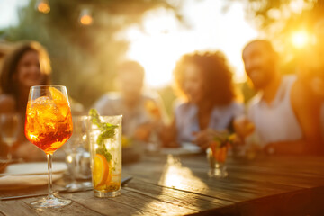 Creative lifestyle concept. Blur background of friends laugh celebrate gather with Aperol Spritz and Mojito cocktail alcohol on focus on wooden patio with gold sunset ray bokeh light. copy text space
