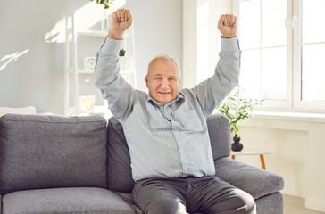 Elderly excited senior man sitting on sofa in living room at home holding hands up in fist like a...
