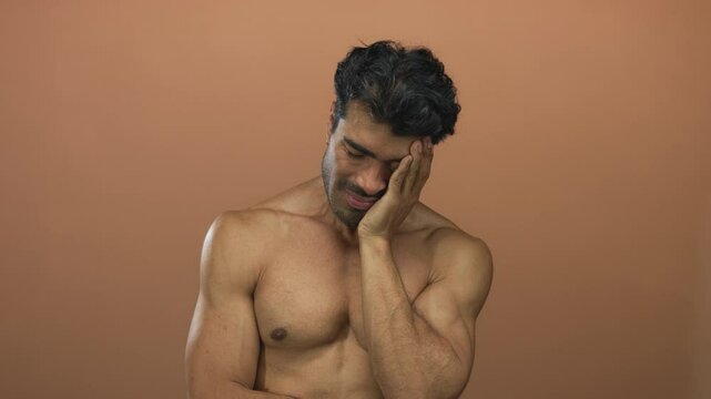 Barechested young hispanic man with hunched posture presses hand to closed eye against brown studio wall; fatigue.