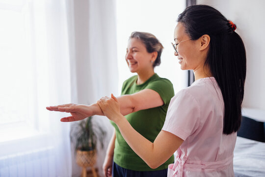 Female physiotherapist assisting a smiling woman in a green shirt with arm exercises in a bright room, showcasing a supportive and encouraging rehabilitation environment