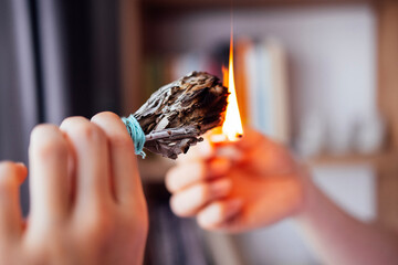 A young woman lighting a bundle of dry aromatic herbs in a room. A girl holds burning incense in her hands against the background of bookshelves and a cozy interior.