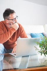 Mature man using laptop at home with mixed expressions. Remote work, digital lifestyle, emotional...