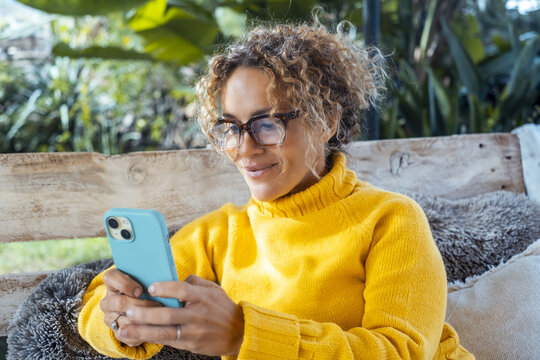 Attractive woman in yellow sweater uses cell phone and relaxes alone in the garden sitting on a sofa. Portrait of serene female writing on cell phone. Mature young people relaxing