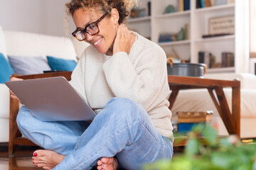 Fototapeta premium Smiling curly-haired woman in her 40s working on a laptop at home. Remote work, online business, modern lifestyle, digital connection and professional independence in a relaxed domestic setting.