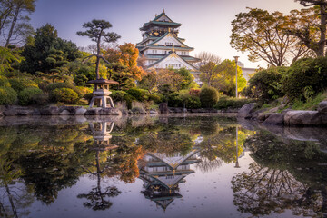 View of a serene pond reflecting the grand Osaka Castle amidst vibrant trees and traditional lanterns, creating a picturesque scene, Osaka, Japan.