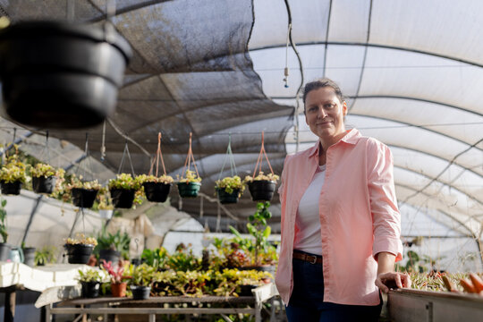 Senior female gardener leaning on bench under shade canopy with plants and hanging pots, copy space - Powered by Adobe