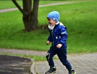 Energetic young boy in blue jacket and hat running with joy on a paved path in green park. Toddler...