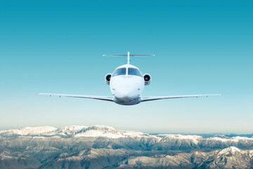 Front view of the white modern luxury executive aircraft flies over snow covered mountains