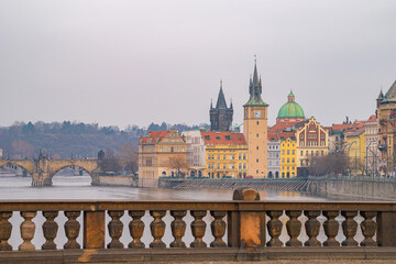 Panoramic view of Prague's Vltava River, featuring a historic bridge, colorful riverfront buildings, and a prominent clock tower under an overcast sky