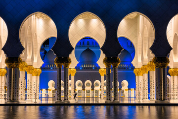 View of illuminated archways and pillars casting reflections on the still water, bathed in blue and gold light, Abu Dhabi, Abu Dhabi, United Arab Emirates.