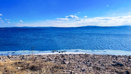 Tranquil blue seascape with a rocky shoreline under a bright sunny sky. Peaceful nature scene of a calm lake or sea with distant mountains and clear water.

