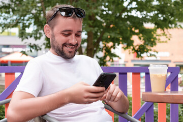 Bearded man with sunglasses on his head enjoying iced coffee while using smartphone at a colorful...