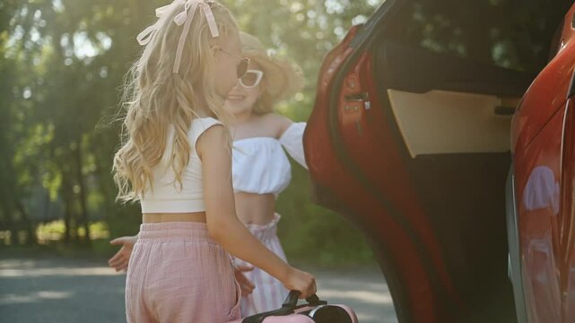 Two little beautiful girls are going to travel together on weekend: caring cheerful kid opening door for cute friend to load suitcase. Smiling children in summer clothes packed luggage for road trip.