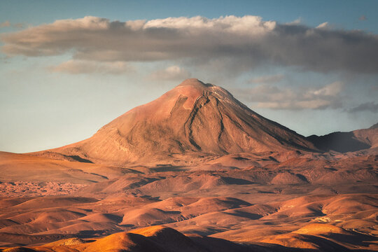 View of a majestic, sun-drenched mountain rises from the ochre desert, its peak softened by a wisp of cloud in the pale blue sky, San Pedro de Atacama, Antofagasta, Chile.