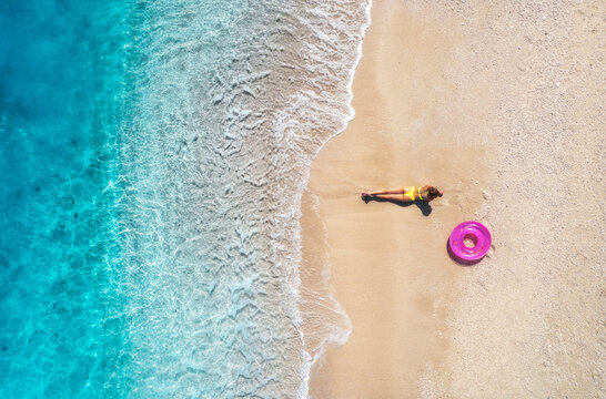 Aerial view with a woman sunbathing on a pristine white sand beach, surrounded by the turquoise waters of Lefkada, Greece, with a pink inflatable ring nearby, embodying summer bliss. Top view of girl