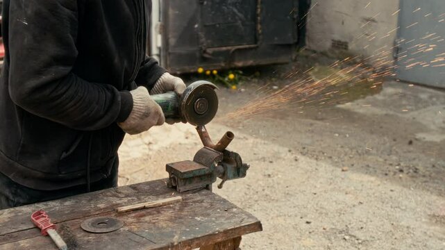 Unrecognizable man in dark jacket cutting metal pipe with angle grinder on cluttered workbench outside